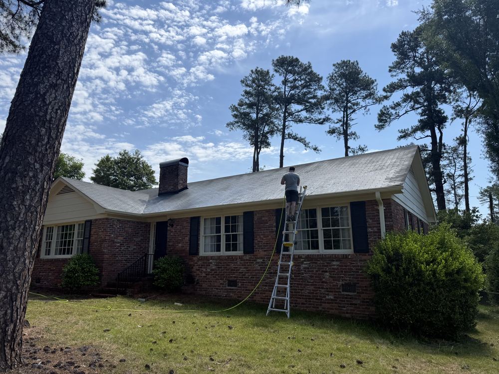 Roof cleaning underway on a long brick ranch home surrounded by pine trees