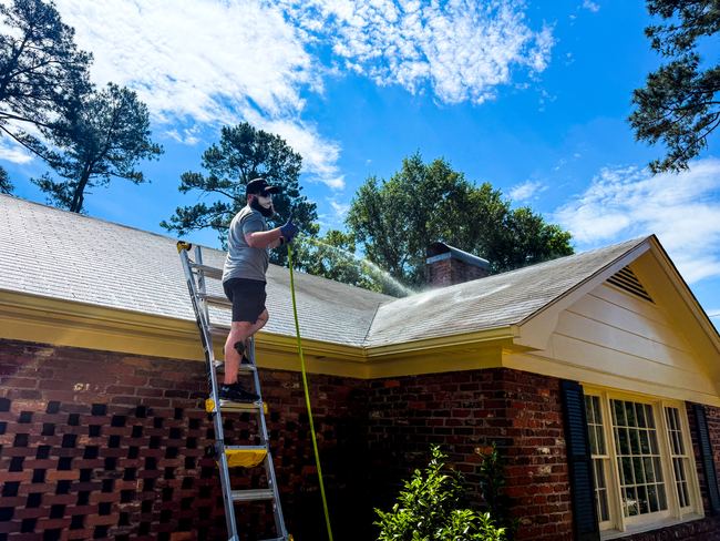 Roof cleaning solution being applied from a ladder toward a roof valley on a brick ranch home
